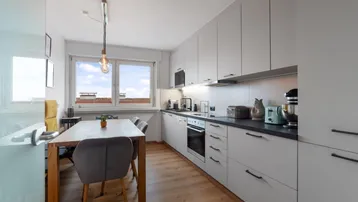 Moderne Einbauküche mit hellgrauen Schränken, dunkler Arbeitsplatte und Essbereich am Fenster.