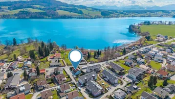Luftaufnahme einer Wohnsiedlung am Seeufer mit Bergen im Hintergrund und markiertem Standort.