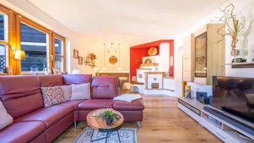 Gemütliches Wohnzimmer mit rotem Ledersofa, Holzboden und traditionellem Kachelofen, hell durch große Fenster.