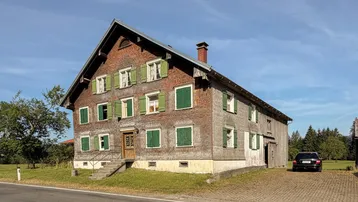 Traditionelles Holzhaus mit grünen Fensterläden in ländlicher Umgebung unter blauem Himmel.