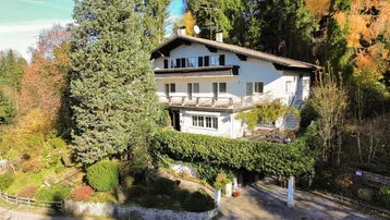 Traditionelles Haus im alpinen Stil, umgeben von üppiger Natur und hohen Bäumen.