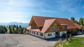 Großes Gebäude mit traditioneller Architektur, umgeben von grünen Hügeln und schneebedeckten Bergen im Hintergrund.