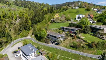 Luftaufnahme einer Wohnsiedlung in hügeliger Landschaft mit viel Grün und verstreuten Häusern.