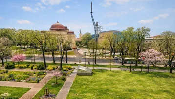 Weitläufige Grünanlage mit Bäumen und historischen Gebäuden im Hintergrund unter blauem Himmel.