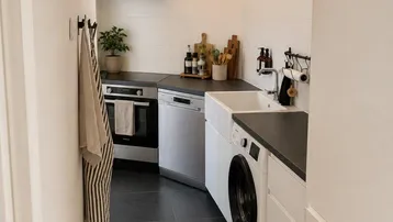 Schmale Küche mit dunklen Fliesen, weißer Einbauküche und integrierter Waschmaschine.