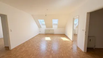 Helles Zimmer im Dachgeschoss mit Parkettboden, Dachschrägen und zwei Dachfenstern, die viel Licht hereinlassen.