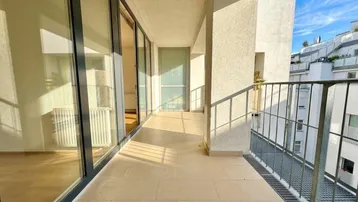Langer, sonniger Balkon mit Fliesenboden und großen Glasfenstern, die viel Tageslicht hereinlassen.