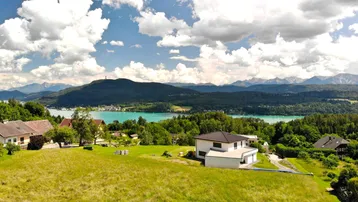 Panoramablick auf ein modernes Haus mit See- und Bergsicht, umgeben von grünen Wiesen und Wäldern.