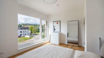 Schlafzimmer mit Holzboden, großem Fenster und moderner Einrichtung, inklusive Ganzkörperspiegel.