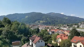 Panoramablick über eine malerische Stadt mit roten Dächern und grünen Bergen im Hintergrund.