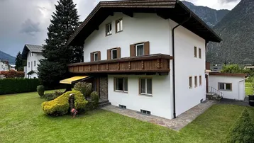 Weißes Haus im alpinen Stil mit Holzbalkon, umgeben von einem grünen Garten und Bergen im Hintergrund.