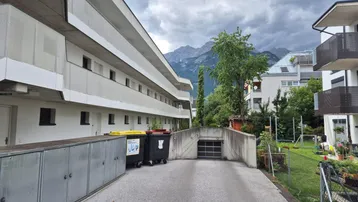 Modernes Mehrfamilienhaus mit Tiefgaragenzufahrt und Bergblick unter bewölktem Himmel.