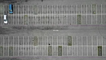Luftaufnahme eines Parkplatzes mit markierten Stellplätzen, zwei Autos und begrünten Zwischenräumen.