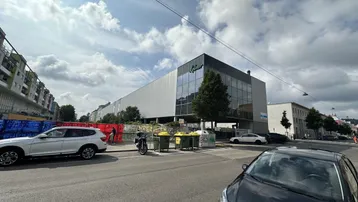 Modernes Geschäftsgebäude mit Glasfassade und Parkplätzen an einer städtischen Straße unter bewölktem Himmel.