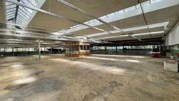 Geräumige, leere Industriehalle mit Betonboden, Oberlichtern und sichtbaren Deckenstrukturen, die Renovierung benötigt.