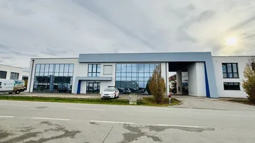 Modernes Geschäftsgebäude mit großer Glasfassade und Parkplätzen davor unter bewölktem Himmel.