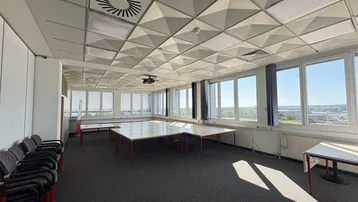Großer Konferenzraum mit vielen Fenstern, modularen Tischen und einem Projektor für Präsentationen.