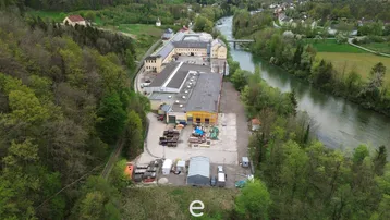 Luftaufnahme eines großen Industrie- oder Gewerbekomplexes am Flussufer, umgeben von viel Grün und Bäumen.