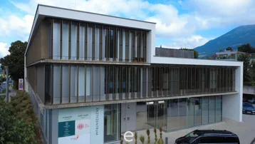 Modernes Ärztezentrum mit großer Glasfassade und Lamellen an der Außenseite, umgeben von Natur und Bergen.