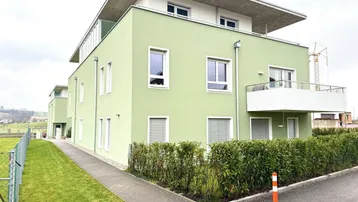 Modernes Mehrfamilienhaus mit hellgrüner Fassade, Balkonen und Flachdach in ländlicher Umgebung.