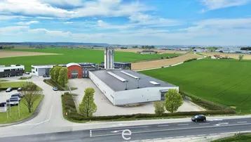 Luftaufnahme eines Gewerbegebiets mit mehreren Gebäuden, umgeben von grünen Feldern unter blauem Himmel.