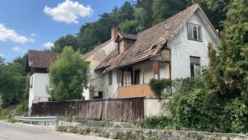Altes, baufälliges Haus mit beschädigtem Dach und Fassade, umgeben von Bäumen und einer Straße.