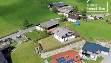 Luftaufnahme eines Hauses mit Grundstück in ländlicher Umgebung, umgeben von grünen Wiesen und Bergen.
