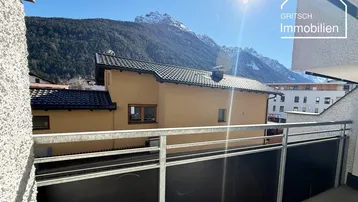Sonniger Balkon mit Blick auf die umliegende Berglandschaft und weitere Gebäude.