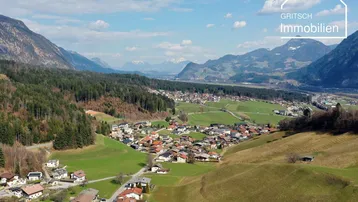 Luftaufnahme eines Dorfes in einem Tal, umgeben von grünen Hügeln und Bergen unter blauem Himmel.