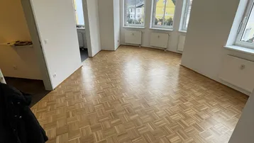 Helles Wohnzimmer mit Fischgrätparkett und großen Erkerfenstern, die viel Tageslicht hereinlassen.