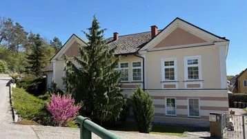 Traditionelles Haus mit heller Fassade, markanten Giebeln und umgebender Grünfläche an einem sonnigen Tag.