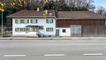 Hausfassade mit angebauter Scheune und grünen Fensterläden an einer Straße.
