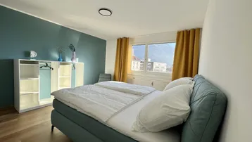 Geräumiges Schlafzimmer mit großem Bett, blauer Akzentwand und Blick auf die Berge.