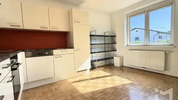 Küche mit hellen Schränken, Parkettboden und einem Fenster, das Tageslicht hereinlässt.