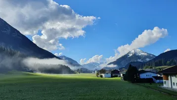 Malerische Alpenlandschaft mit grünen Wiesen, schneebedeckten Bergen und morgendlichem Nebel in einem Tal.