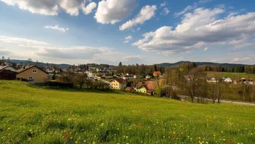 Weitläufige Landschaft mit einem malerischen Dorf, umgeben von grünen Wiesen und sanften Hügeln unter blauem Himmel.