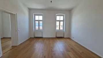 Helles, leeres Zimmer mit Holzboden und zwei Fenstern, bereit zur individuellen Gestaltung.