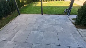 Geräumige, geflieste Terrasse unter einer Überdachung, umgeben von einer grünen Hecke.