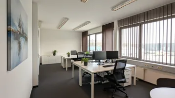 Helles und modernes Großraumbüro mit mehreren Arbeitsplätzen, großen Fenstern und Teppichboden.