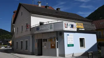 Gebäudeansicht mit hellem Anstrich, kleinem Balkon und Geschäftsräumen im Erdgeschoss.