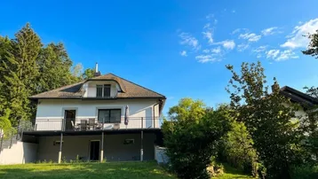 Freistehendes Haus mit Balkon und Garten auf einem grünen Hügel unter blauem Himmel.