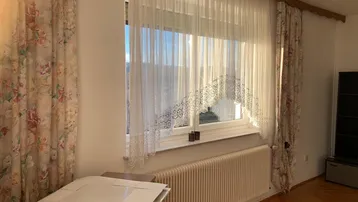 Helles Zimmer mit floralen Vorhängen, Parkettboden und einem Fenster, das viel Sonnenlicht hereinlässt.
