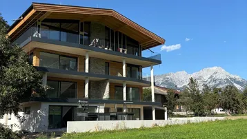 Modernes Apartmentgebäude mit Holz- und Steinfassade, großen Balkonen und Bergblick unter blauem Himmel.