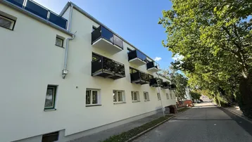 Moderne Wohnhausfassade mit Balkonen und großen Fenstern, umgeben von grünen Bäumen.