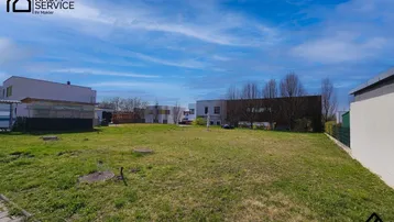 Leeres Grundstück mit Grasfläche und umliegenden Gebäuden unter blauem Himmel.
