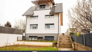Modernes Mehrfamilienhaus mit weißer Fassade, dunklem Dach und kleinem Vorgartenbereich.