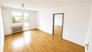 Helles Wohnzimmer mit Parkettboden, großem Fenster und Zugang zu einem Balkon.