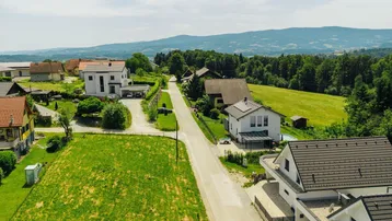 Luftaufnahme einer ländlichen Wohnsiedlung mit Häusern, grünen Feldern und bewaldeten Hügeln im Hintergrund.
