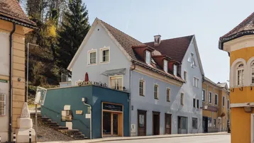 Historisches Gebäude mit hellblauer Fassade, rotem Ziegeldach und einer kleinen Terrasse an der Straße.