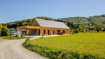 Neubauhaus in Holzbauweise mit Satteldach, umgeben von grünen Wiesen und Hügeln unter blauem Himmel.